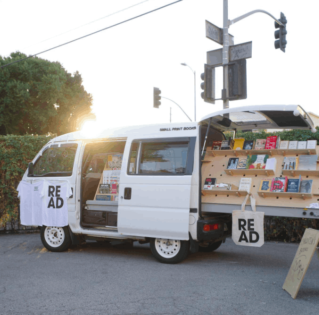 White compact van with door ajar and bookcase coming out of its rear, parked outside against a green hedge in a parking lot with the sun setting behind it.